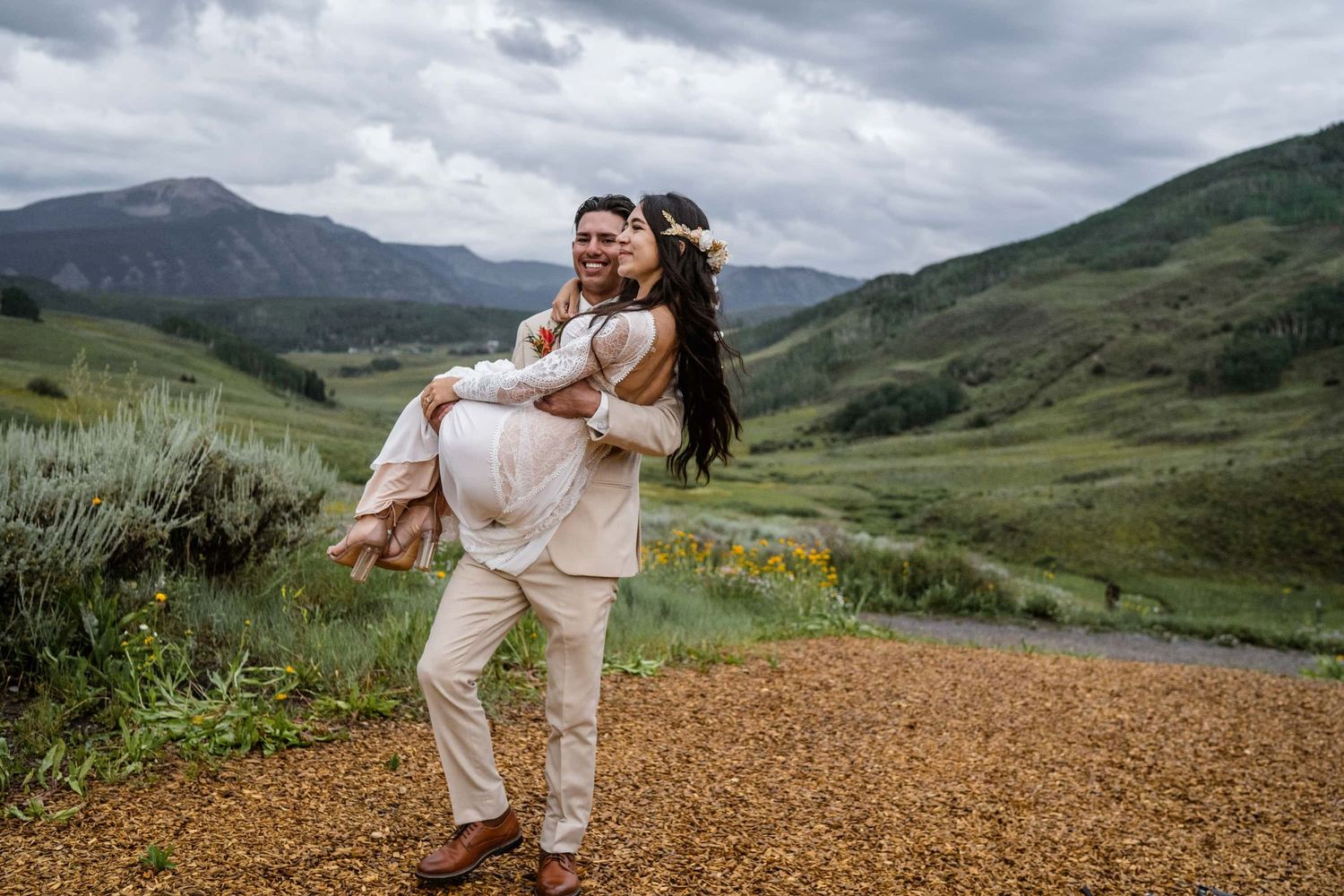 A couple embraces on a mountain trail with dramatic cloudy skies and green rolling hills in the background.
