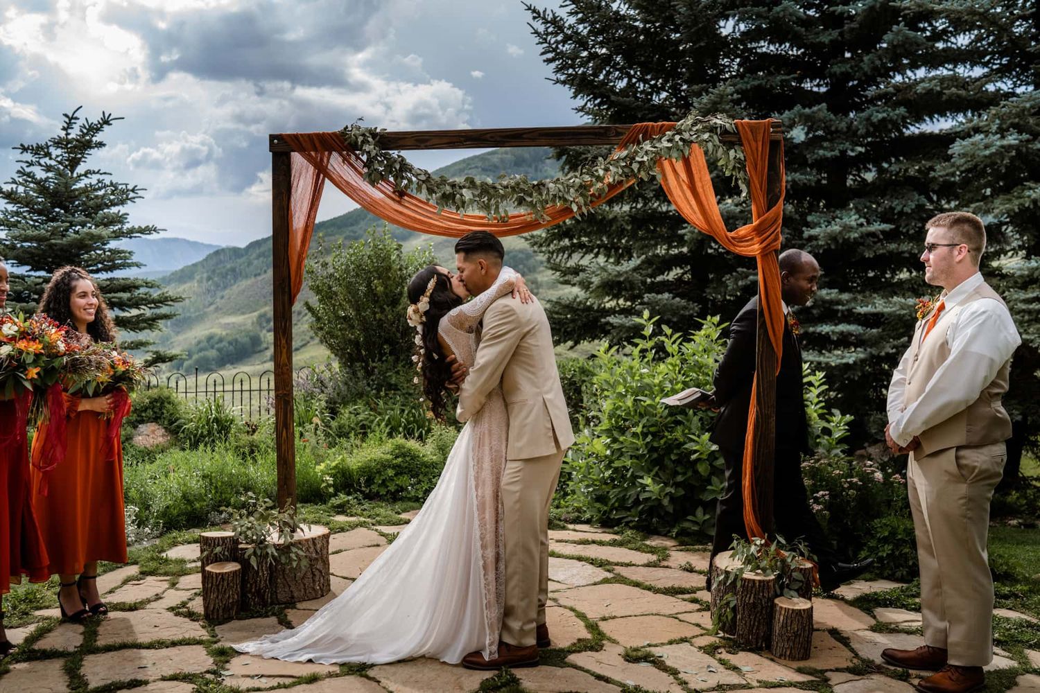 A scenic mountain wedding ceremony takes place under a wooden arch decorated with orange fabric and greenery.