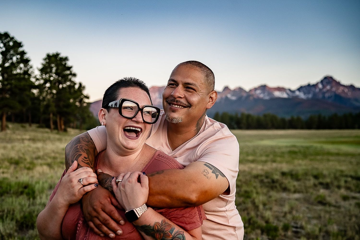 A couple shares a joyful moment in a meadow with mountains in the background at sunset.