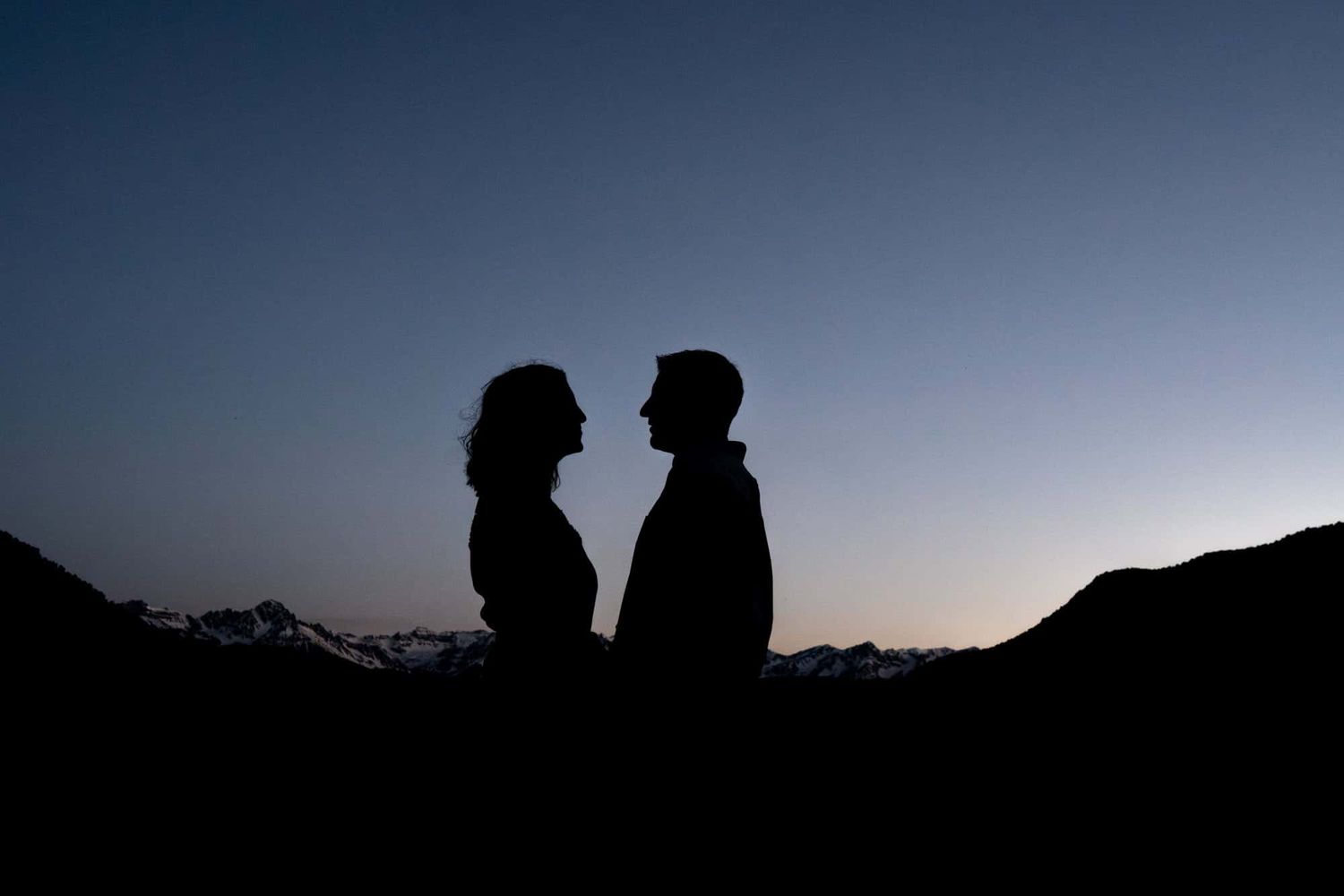Silhouette of two people standing close together against a dusky mountain backdrop.