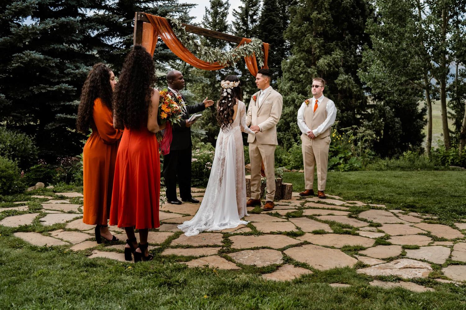 Outdoor wedding ceremony taking place on stone pathway with rustic wooden arch decorated with orange fabric and greenery.