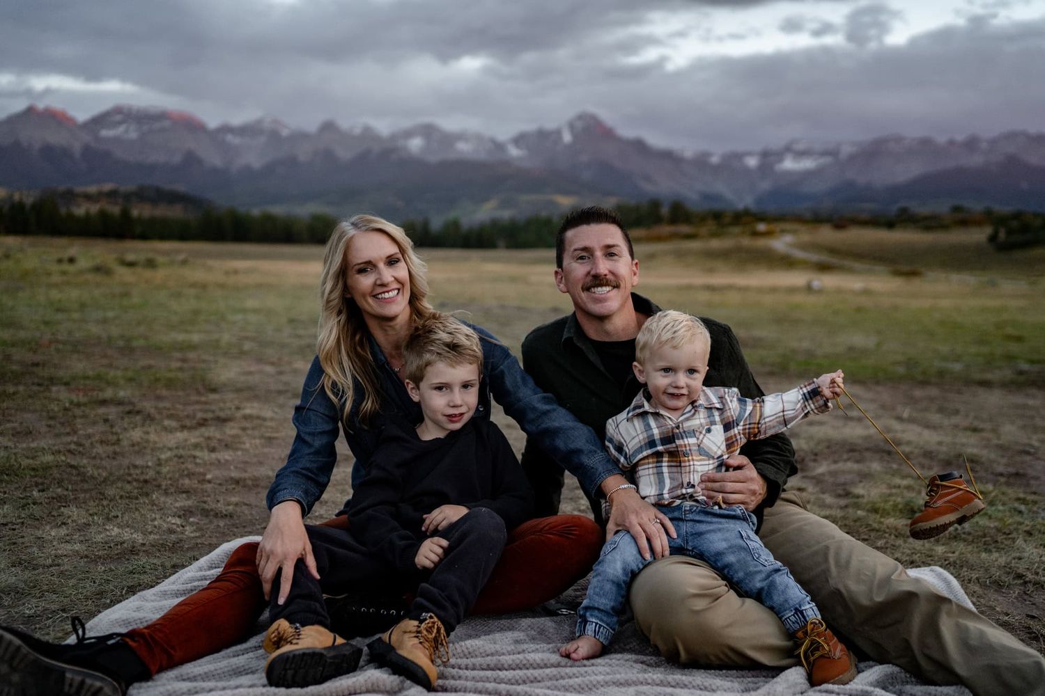 Family sits together on blanket laughing with mountain range visible in background at dusk.