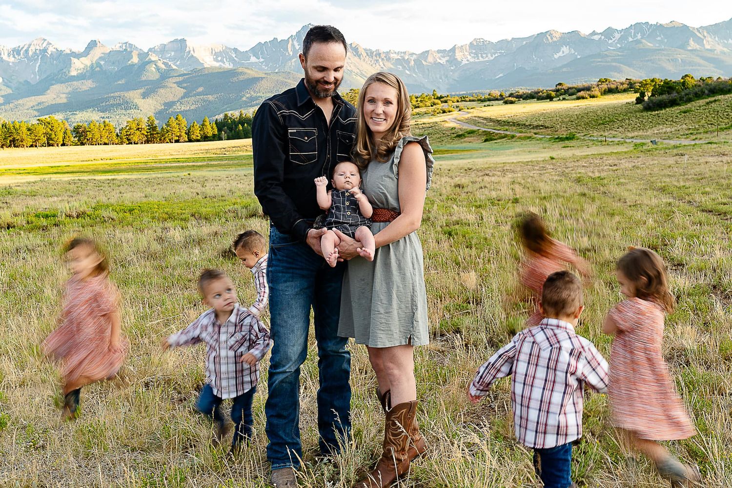 Family portrait in mountain field with children running and playing in background.