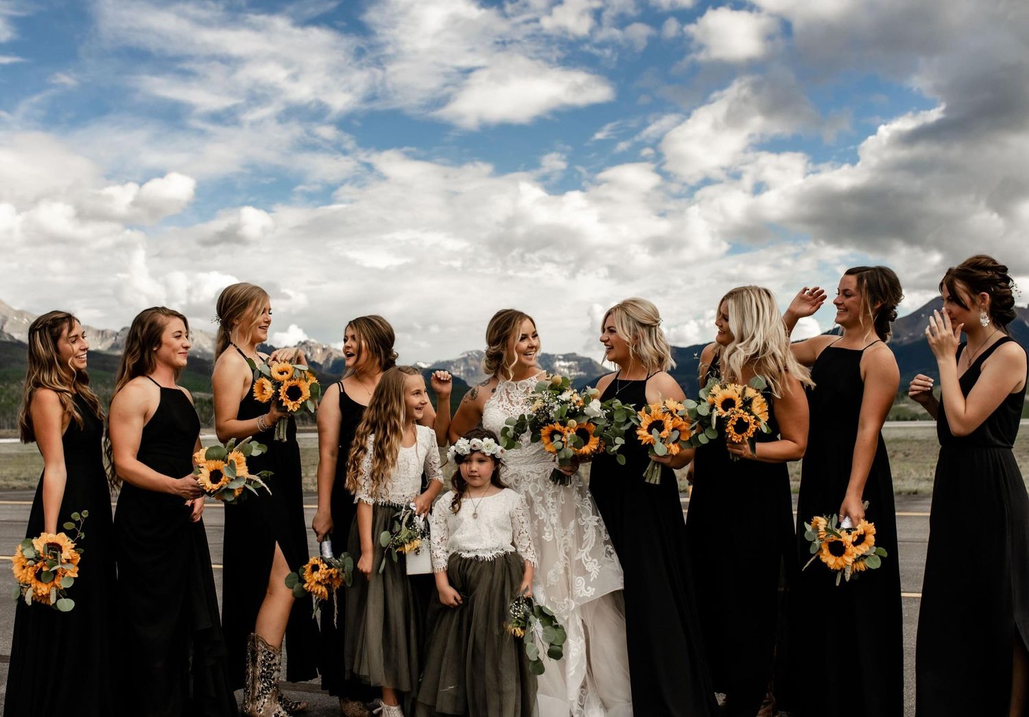 Wedding party gathered outdoors under dramatic cloudy sky holding bright sunflower bouquets.