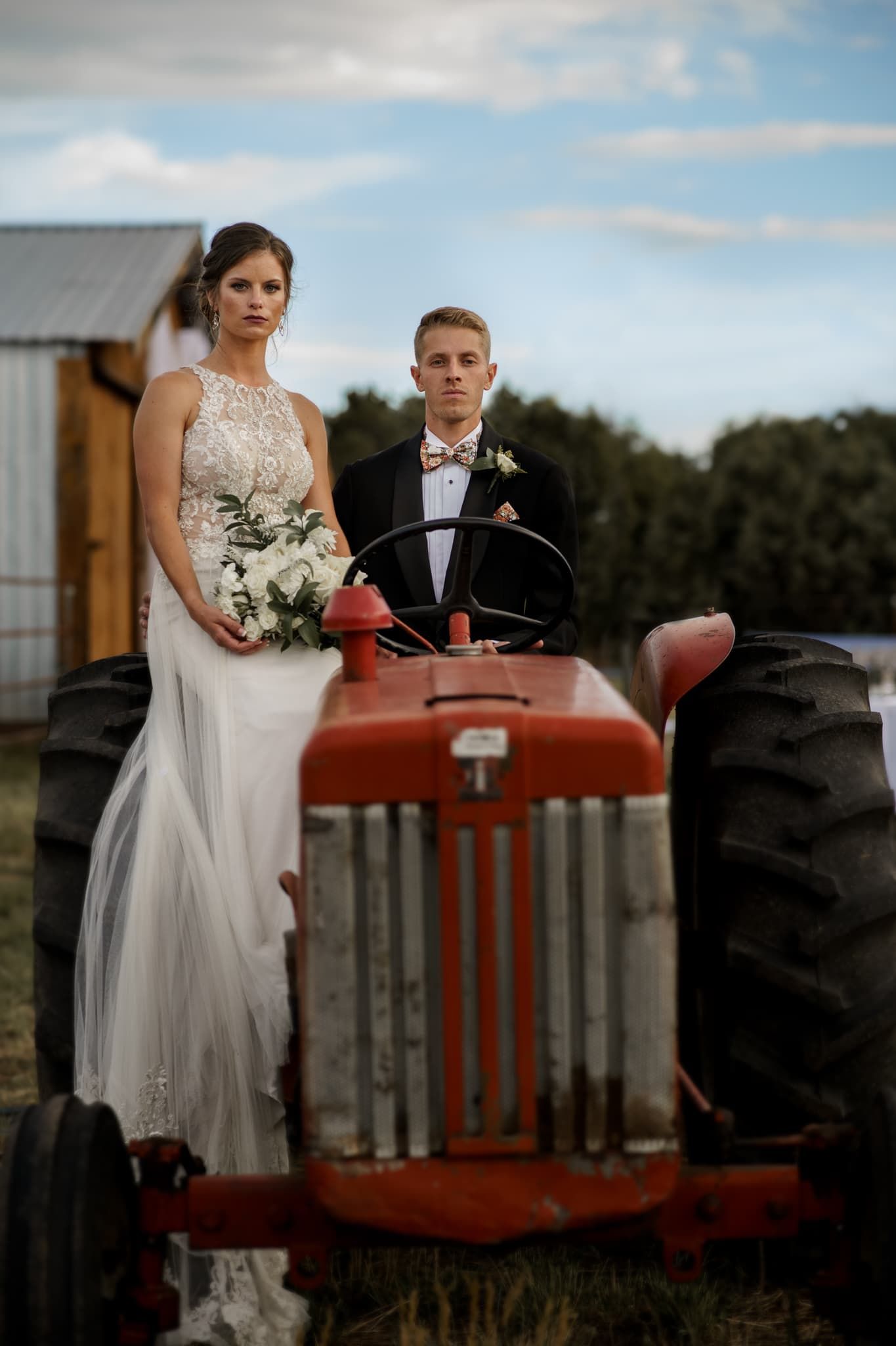 A newlywed couple poses with a vintage red tractor on their rustic farm wedding day.