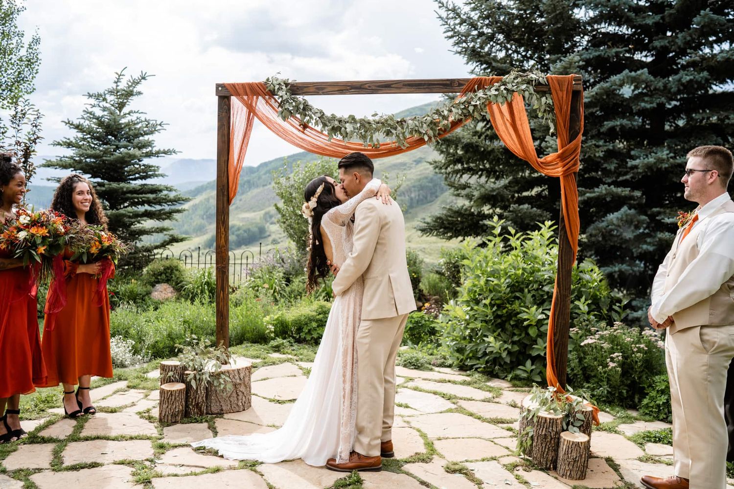 An outdoor mountain wedding ceremony takes place under a wooden arch decorated with orange and white fabric and flowers.