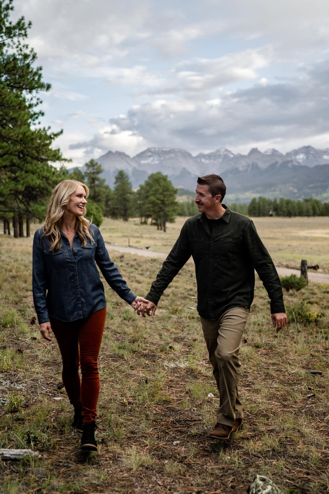 A couple walks hand in hand through a mountain meadow with snow-capped peaks in the background.
