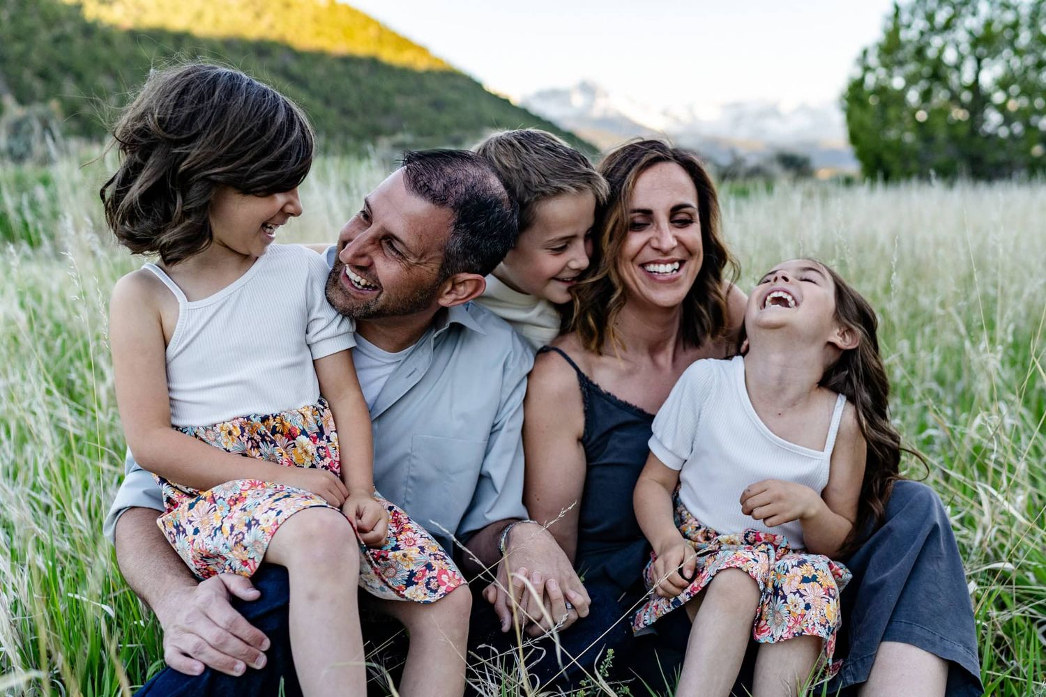 Family shares joyful laughter together while sitting in meadow at sunset. Family photographer in Montrose Colorado met them to do this.