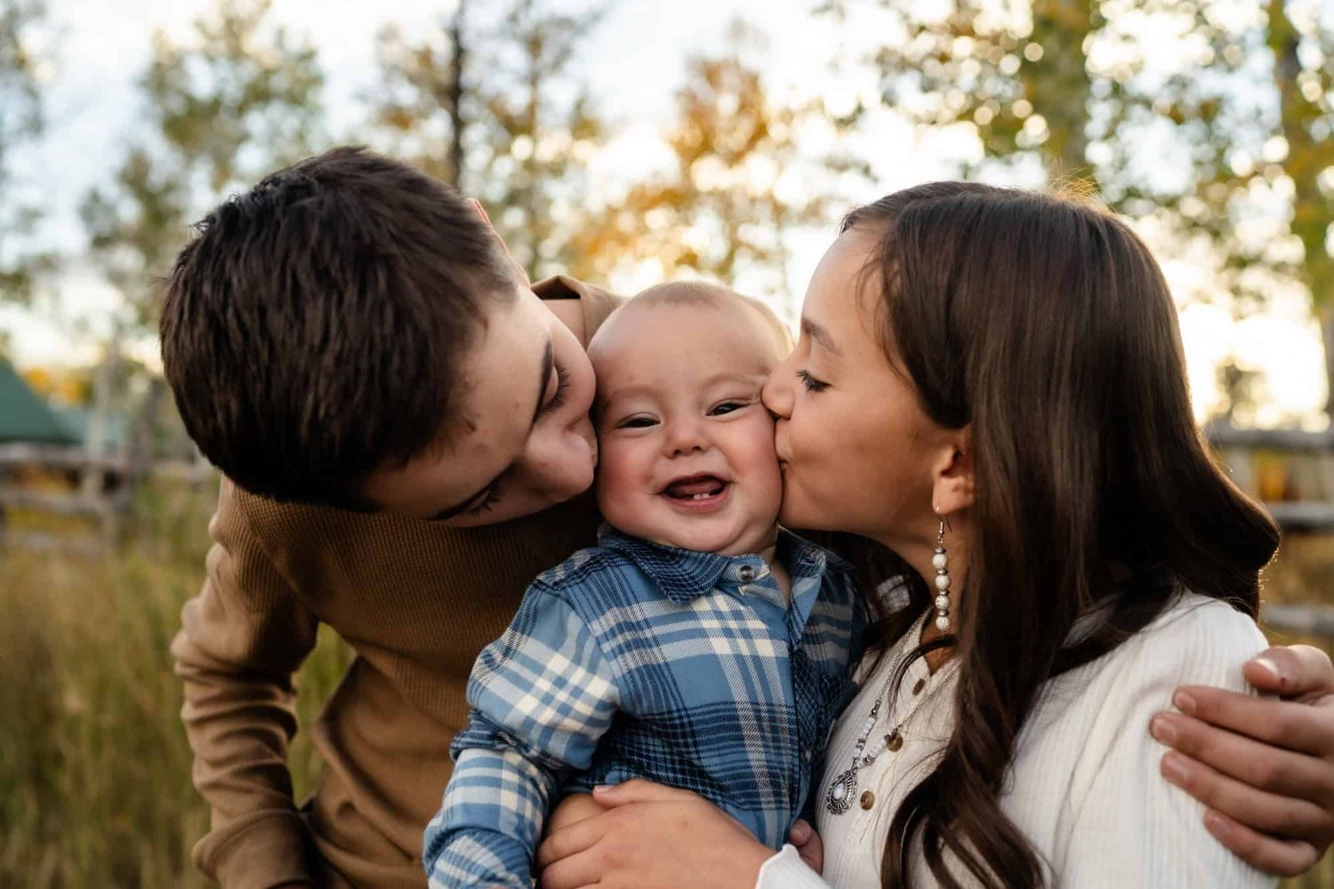 Baby laughing with joy while receiving kisses outdoors during golden hour in a natural setting.