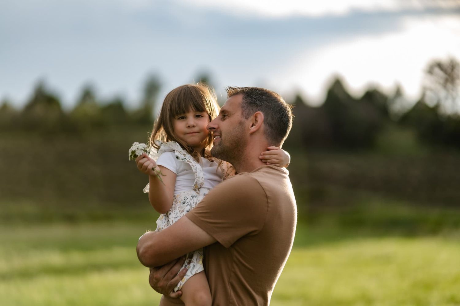 A parent holds a small child in a floral dress against a backdrop of rolling hills.