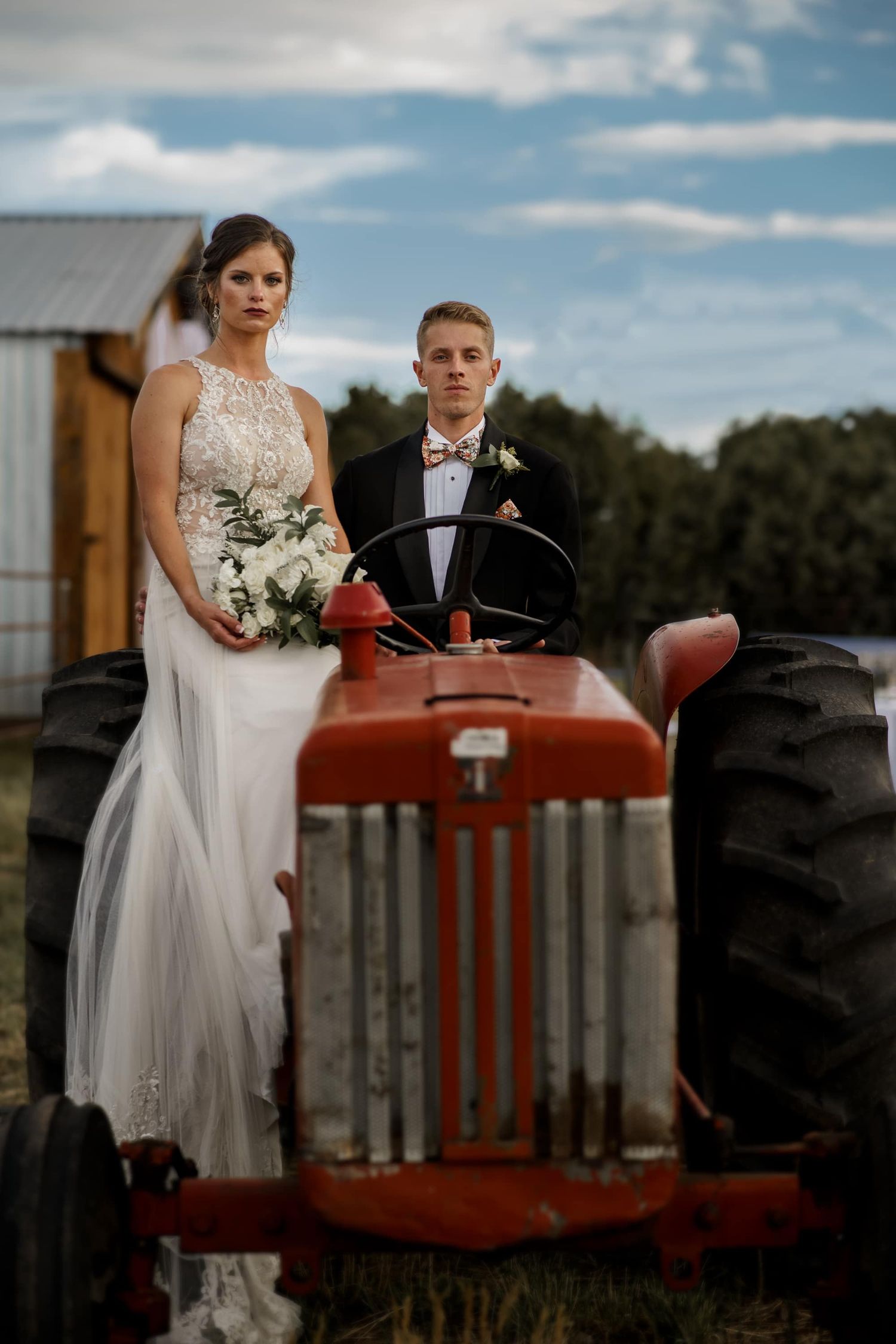 Newlyweds pose on vintage red tractor against rustic farm backdrop.