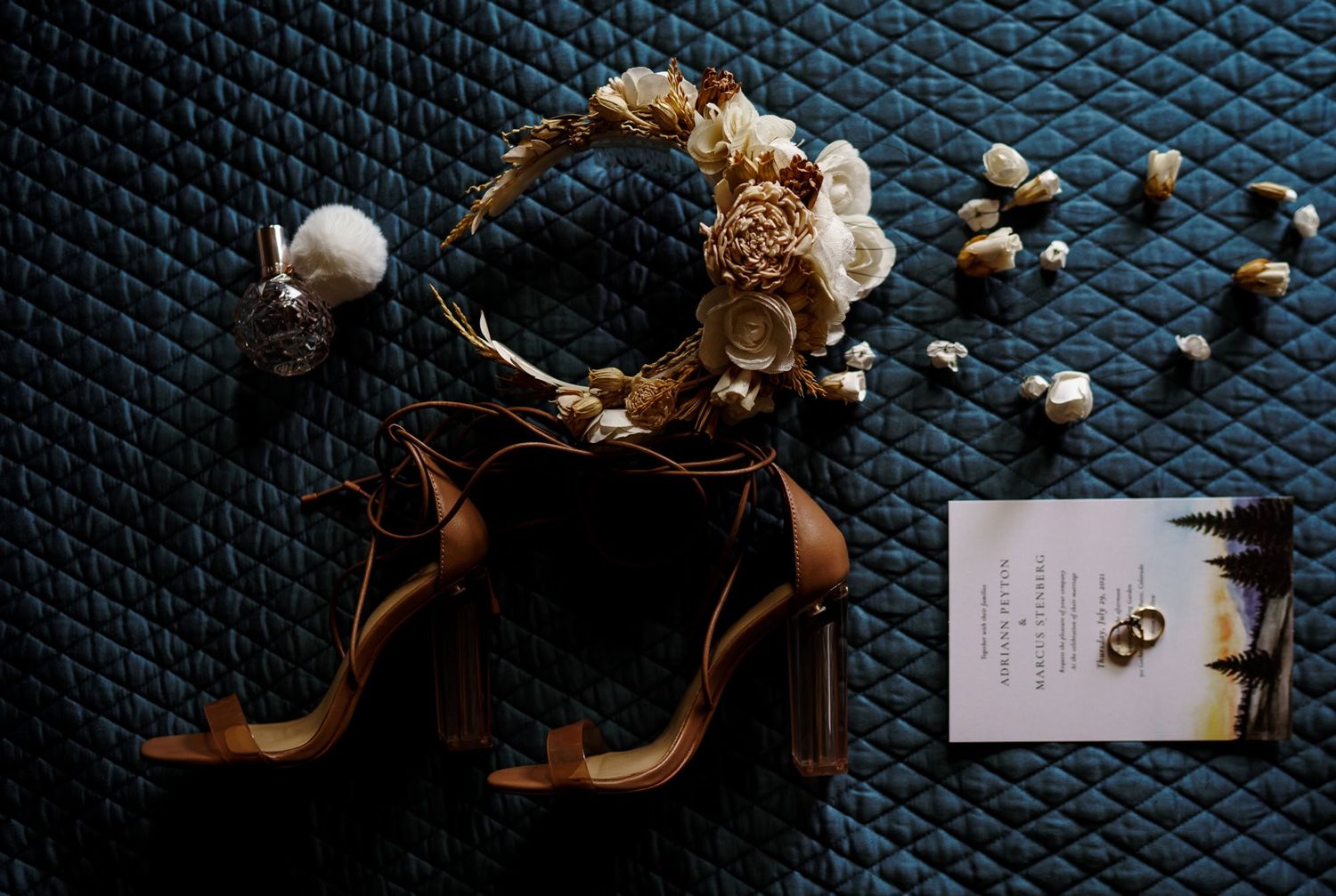 Brown bridal heels and dried floral arrangements displayed on dark textured surface with wedding invitation.