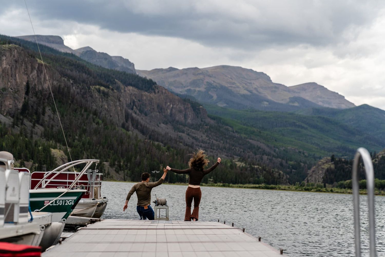 Friends jump joyfully on a wooden dock with scenic mountain landscape and lake views.