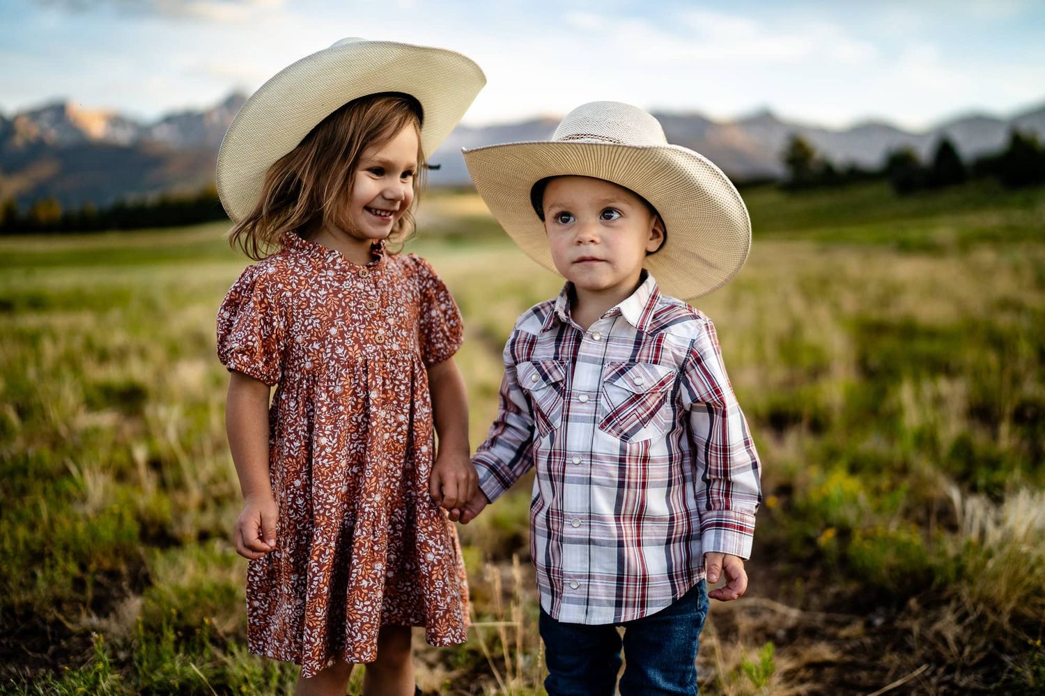 Young siblings in western wear stand together in a mountain meadow at sunset.