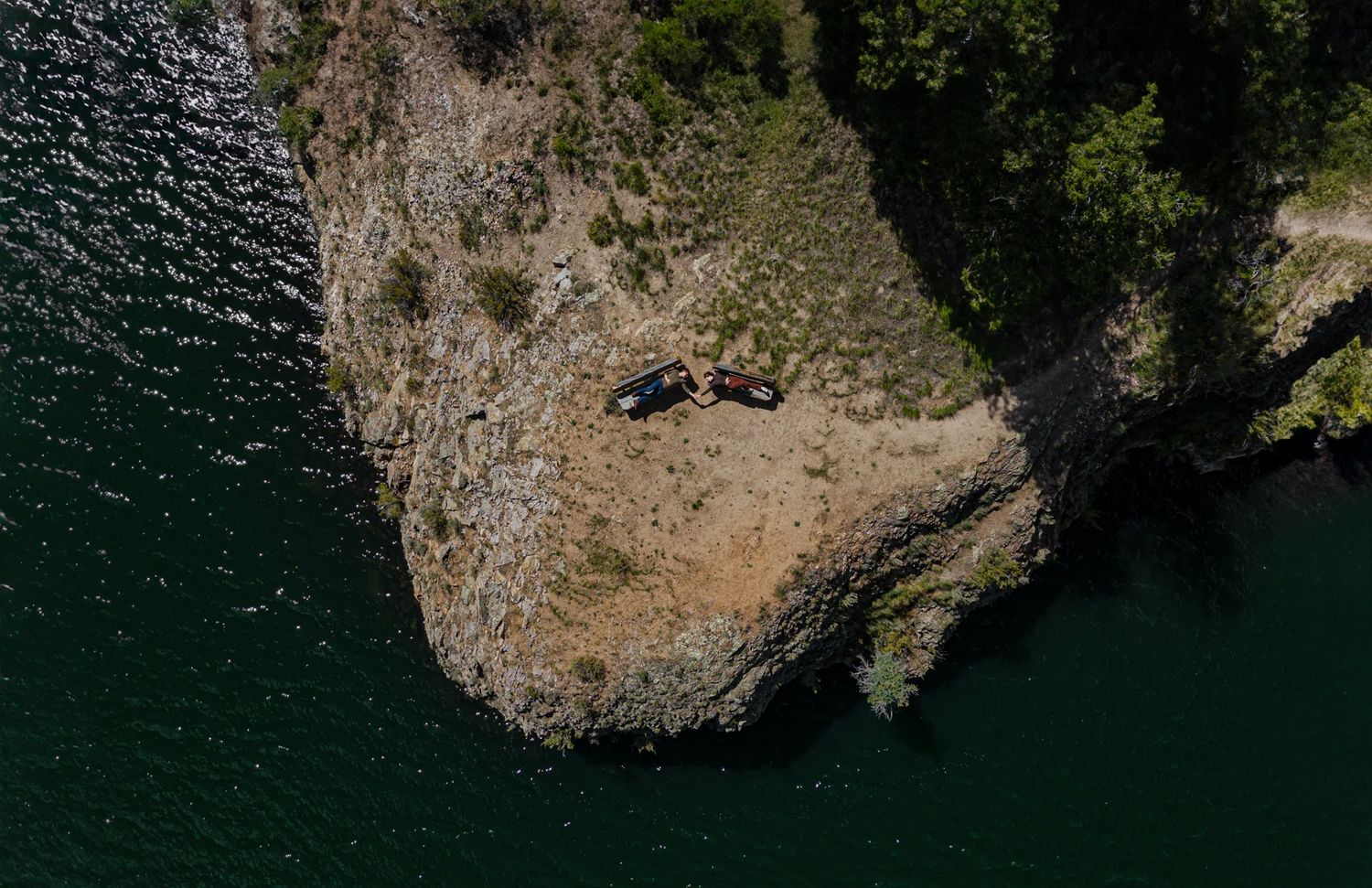Aerial view of a couple lying on a rocky cliff edge overlooking dark green waters. Captured by Lake City, Colorado photographers