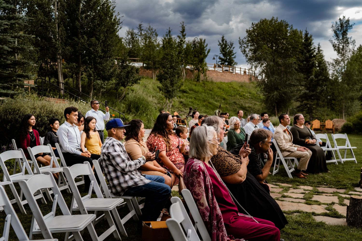 Wedding guests seated in white chairs at an outdoor ceremony venue with evergreen trees and dramatic cloudy sky.