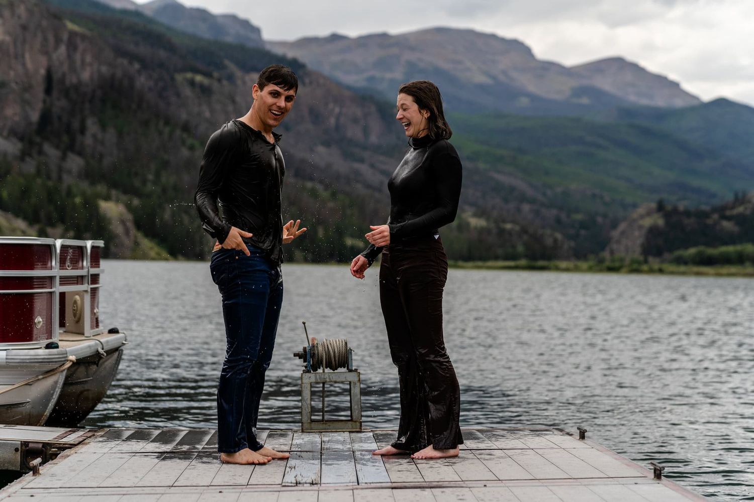 Two people standing on a wooden dock having a conversation with mountains in the background.