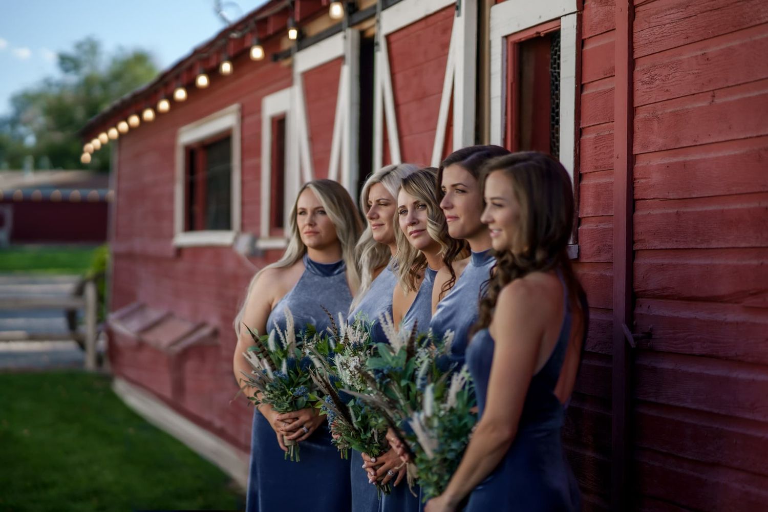 Bridesmaids in navy blue dresses stand together holding bouquets against a rustic red barn venue near Montrose Colorado.