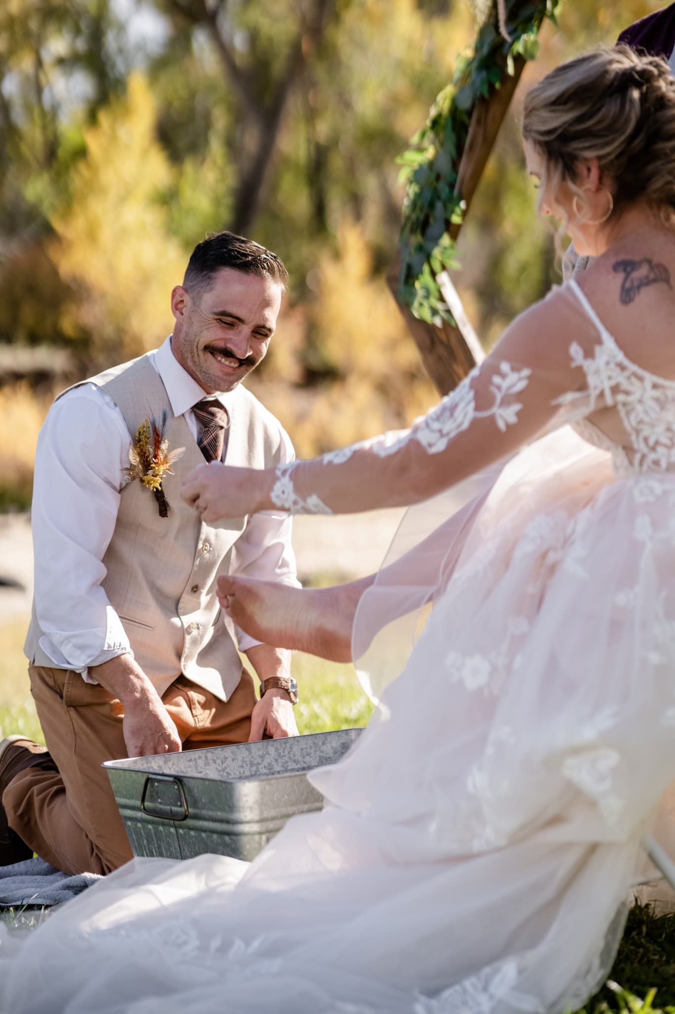Groom in light vest kneels while laughing during outdoor wedding ceremony moment.