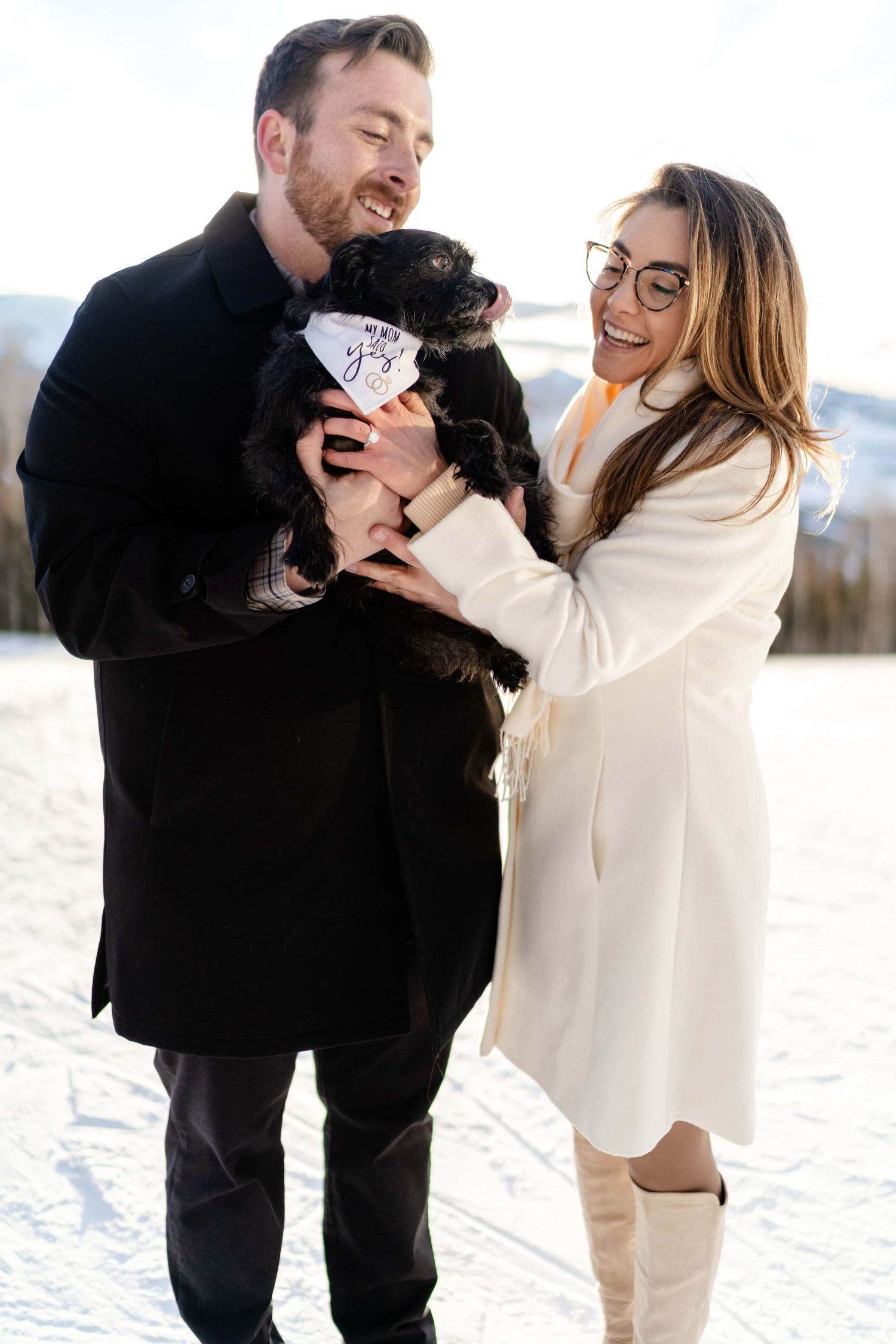 Happy couple in winter coats holding their small black dog in a snowy mountain setting.