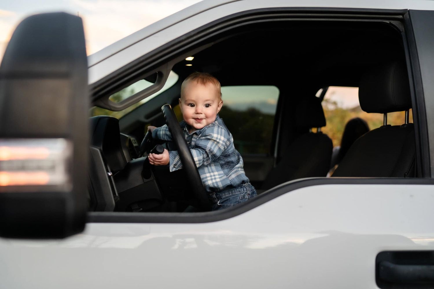 Smiling toddler sitting in driver's seat of parked car looking through window.