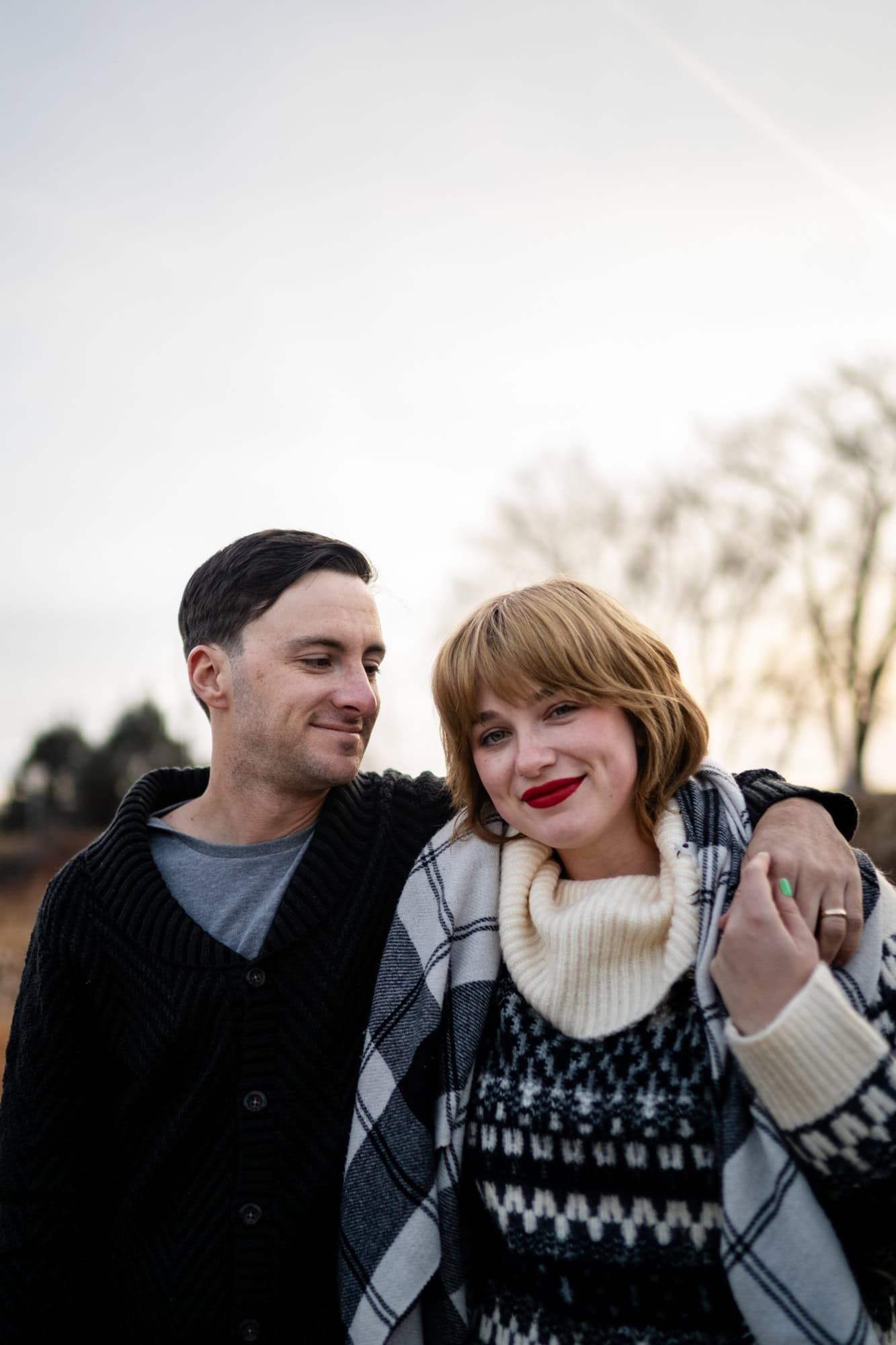 Couple wearing warm winter sweaters embrace and smile outdoors on a cloudy day.