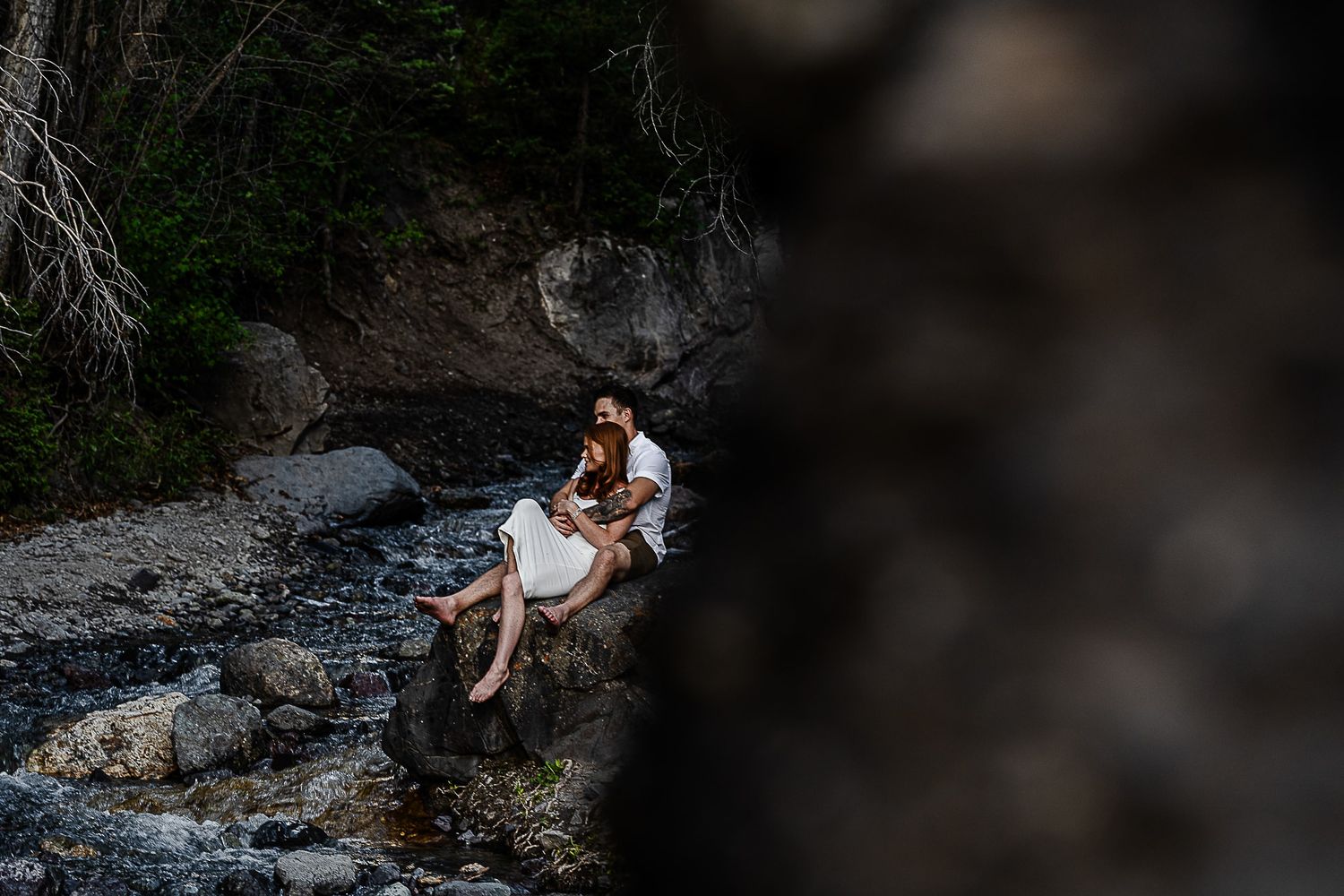 Couple embracing on rocks by mountain stream in romantic wilderness photo session.