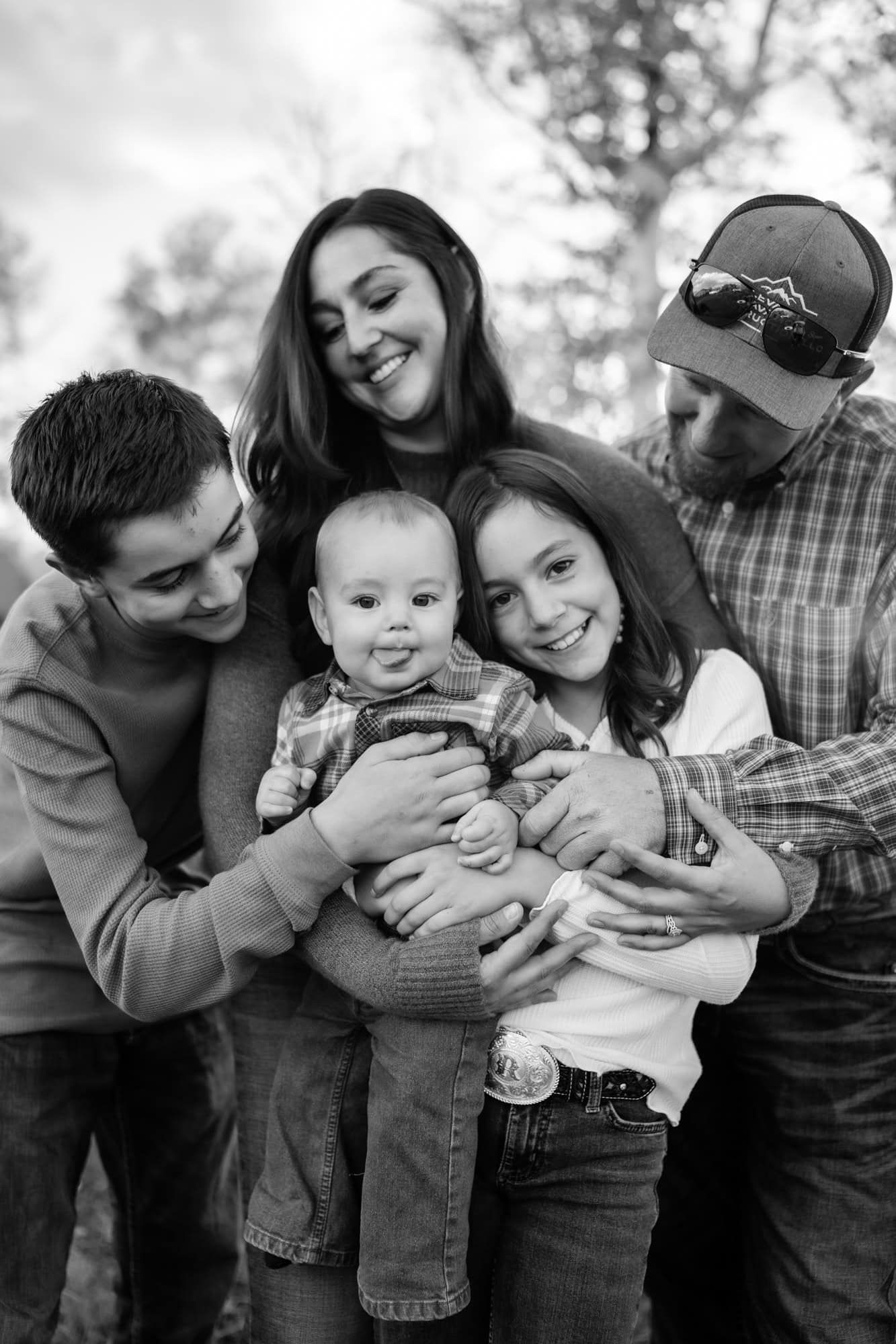 A joyful family portrait featuring a baby in denim being held and surrounded by loved ones in an outdoor setting.