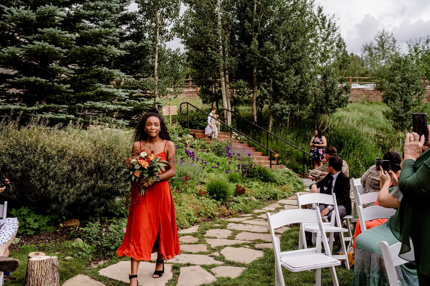 Someone in a vibrant red dress walks down a garden path during an outdoor wedding ceremony as guests watch and take photos.