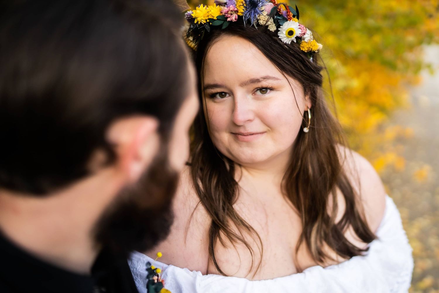 Bride wearing floral crown looks lovingly at partner during autumn wedding.