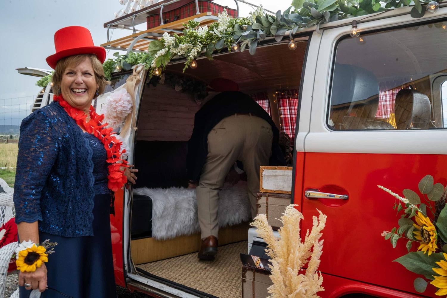 Person in red hat standing by decorated vintage red VW van with flowers and greenery.