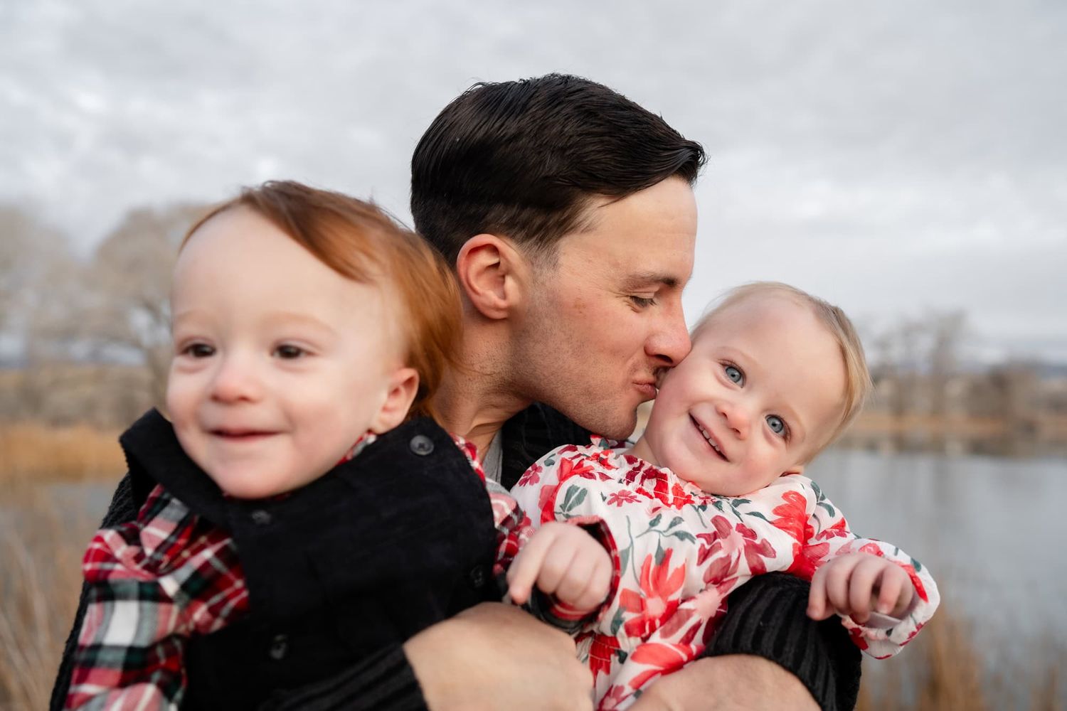 A parent shares a kiss with two smiling babies during an outdoor photoshoot.