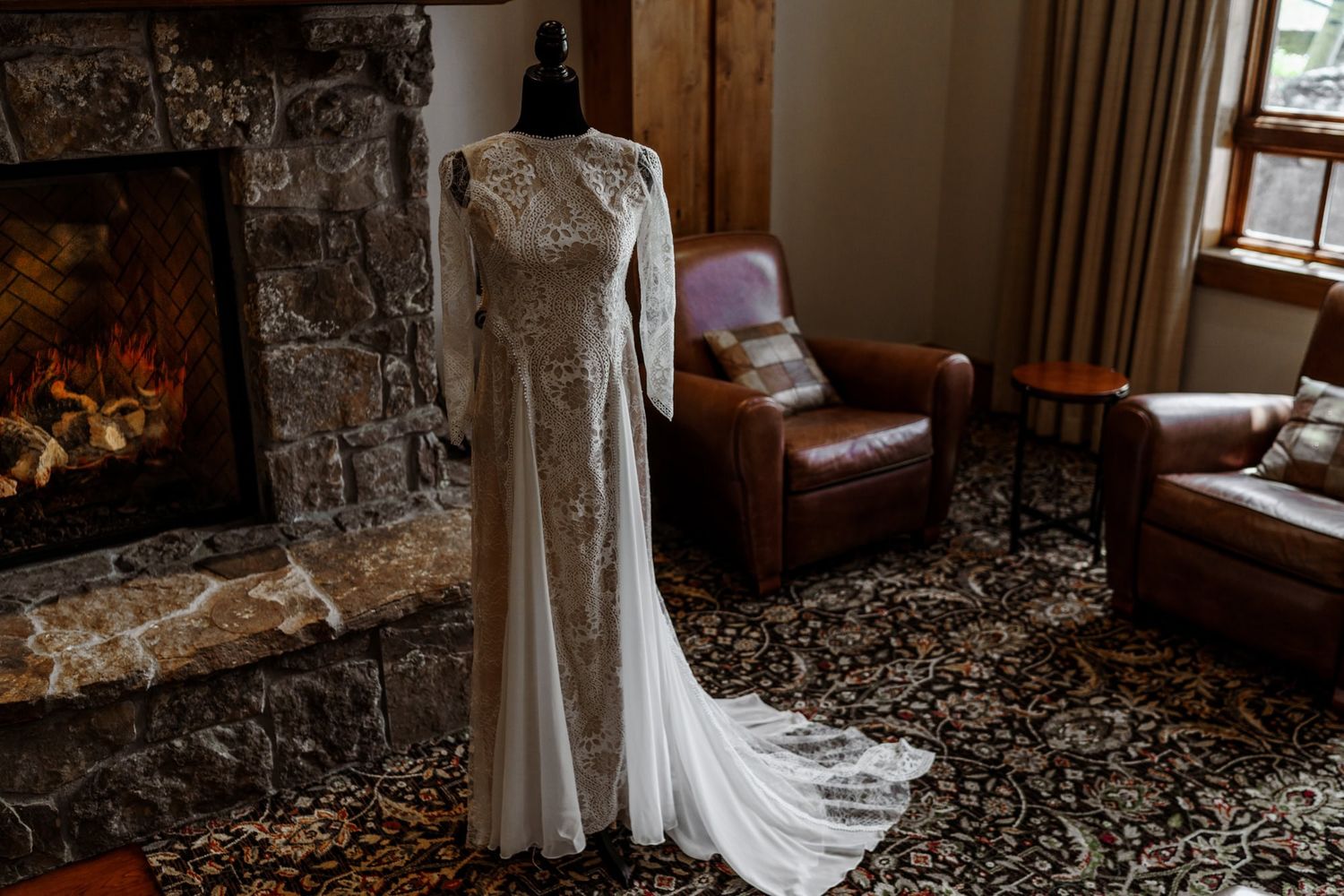A flowing white lace wedding dress hangs in front of a stone fireplace with leather chairs in a rustic room.