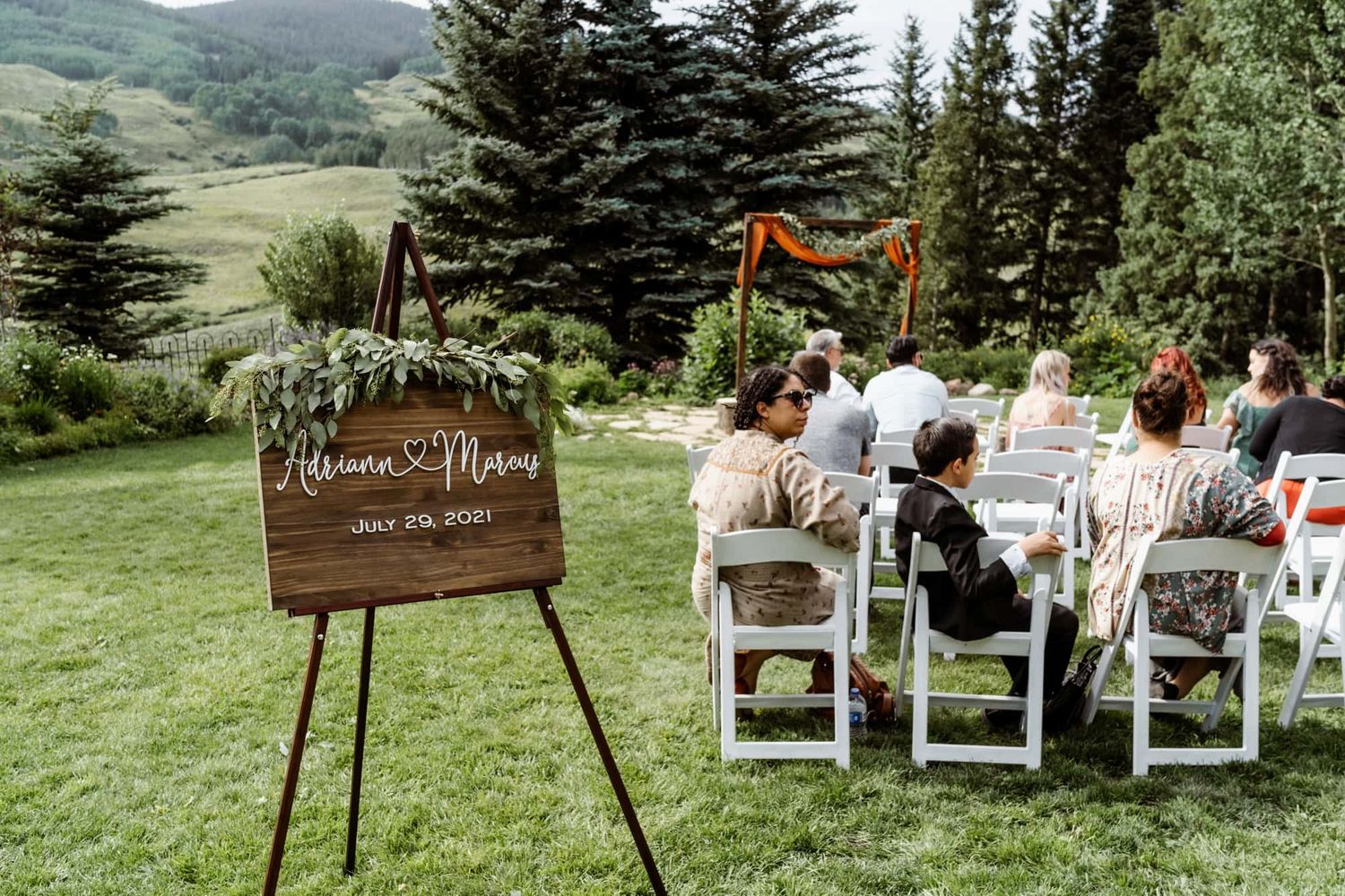 A wooden welcome sign stands at an outdoor mountain wedding ceremony with white chairs and evergreen trees.