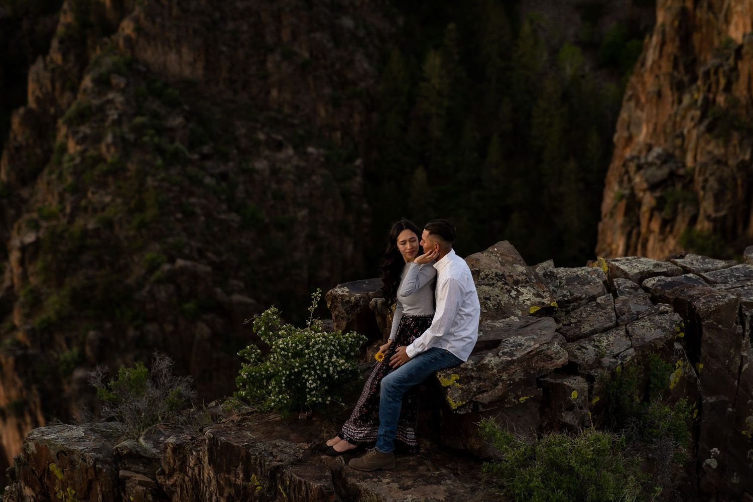Couple sitting on rocky cliff edge with dramatic mountain vista behind them.