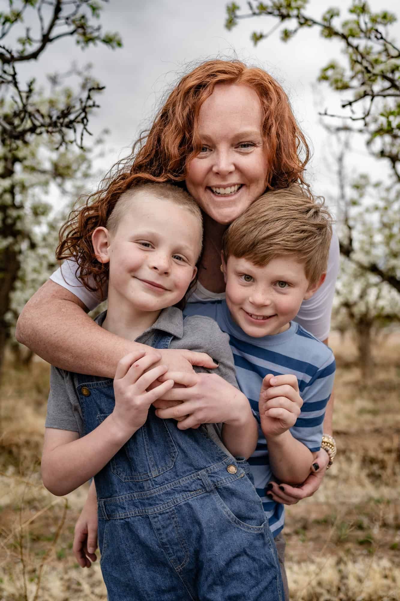 Happy family portrait of three people hugging and smiling outdoors among trees.
