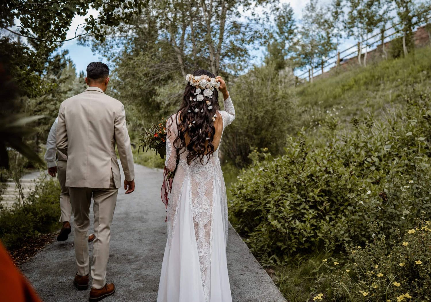 Two people walk down a scenic forest path, with one wearing a flowing white lace dress and floral crown.