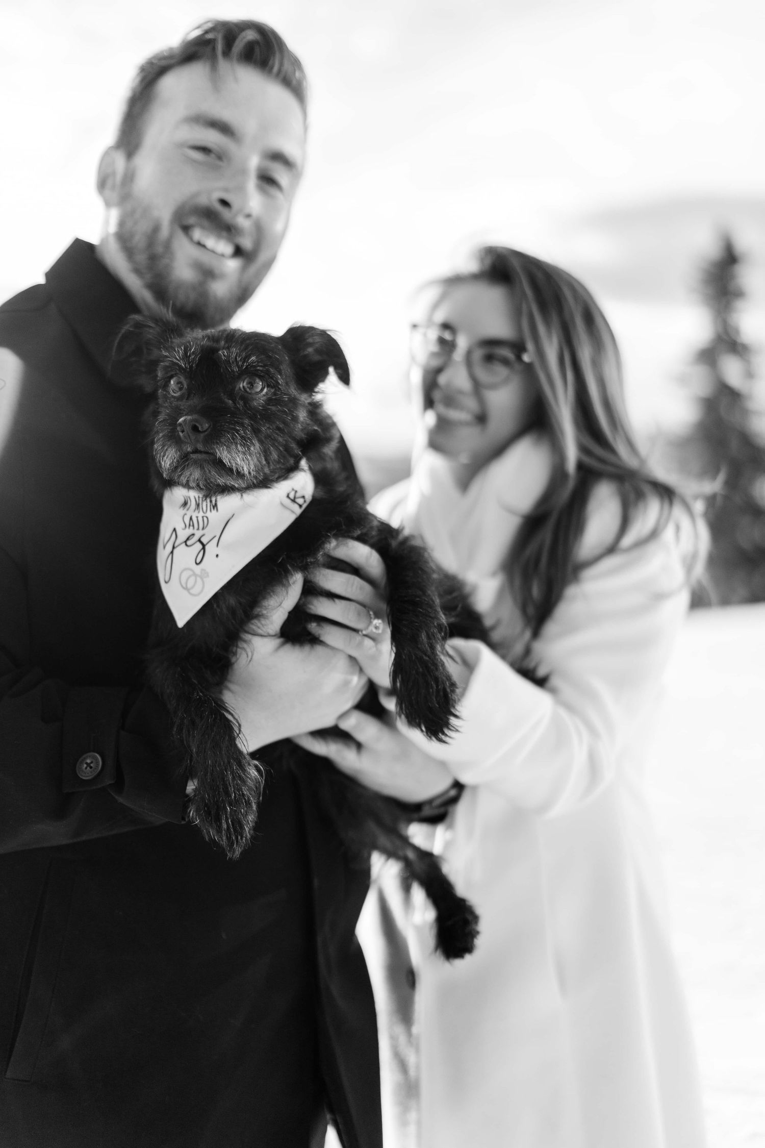 Black and white photo of a couple with their small dog wearing a bandana in the snow.