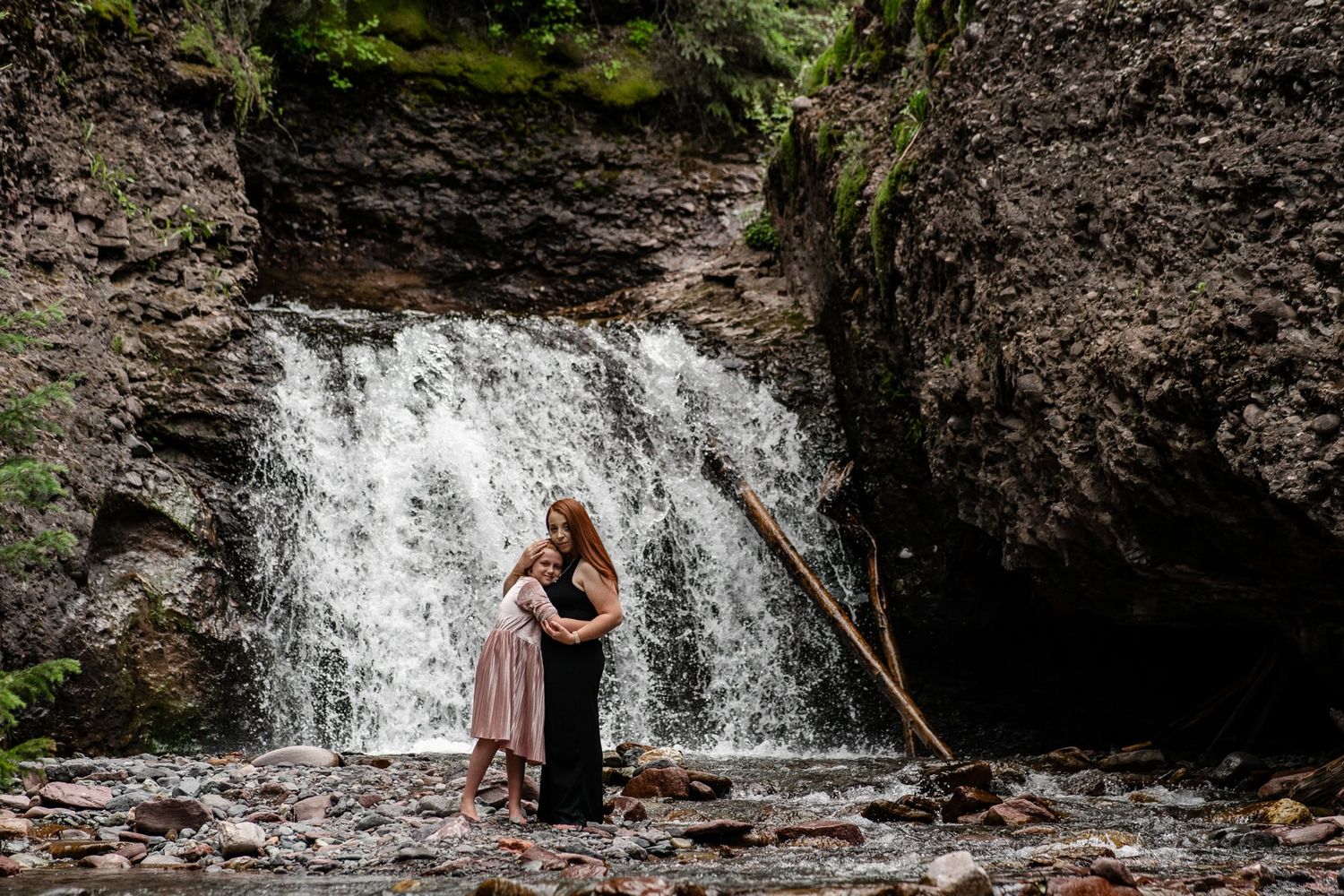 Mom hugs daughter in flowing dress standing before majestic waterfall in Telluride Colorado.