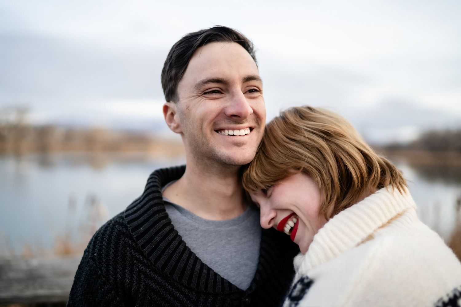 Happy couple in winter attire laughing together by a lake.