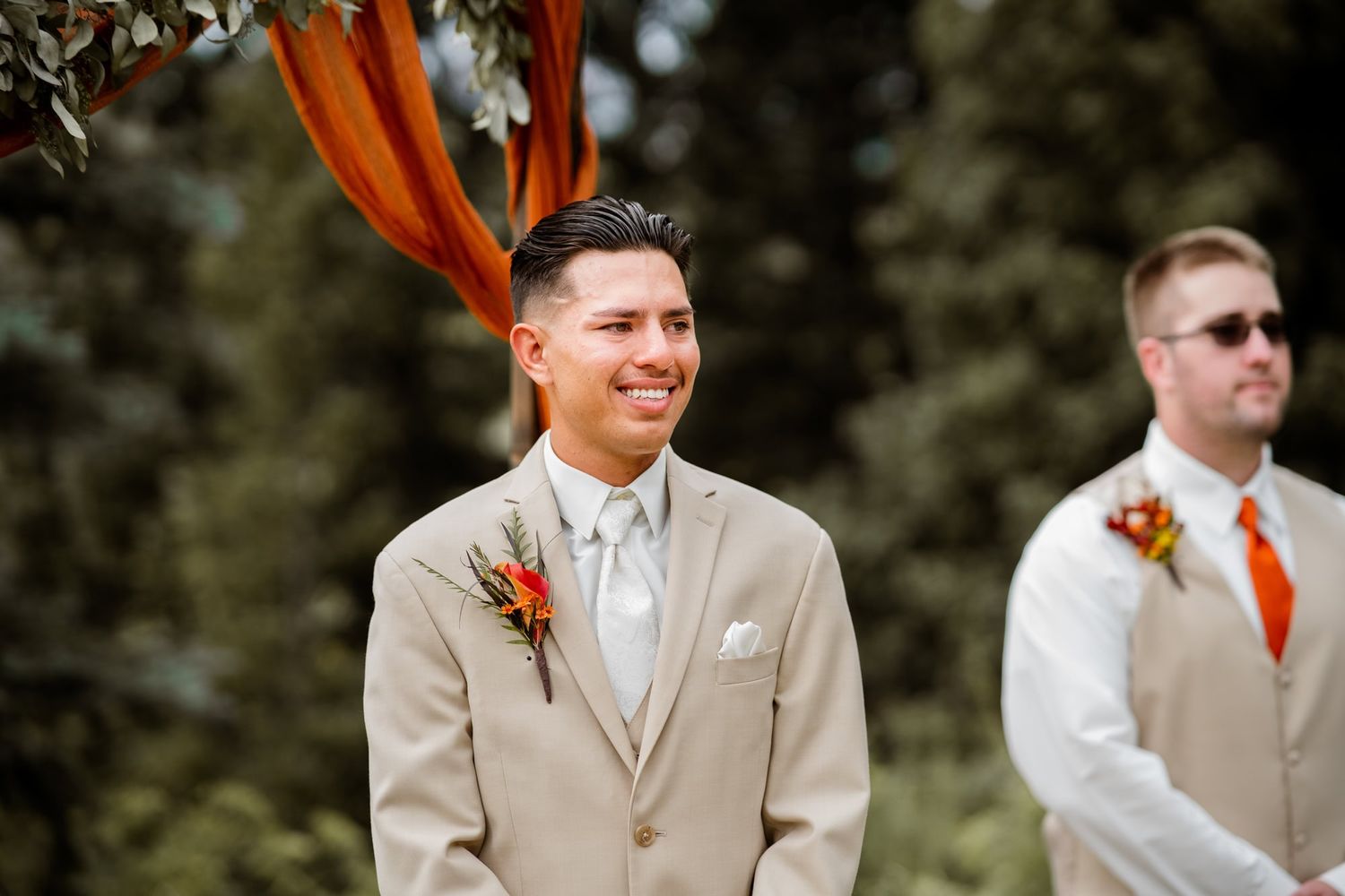 A groom in a tan suit with orange boutonniere smiles during an outdoor wedding ceremony while groomsman stands behind.