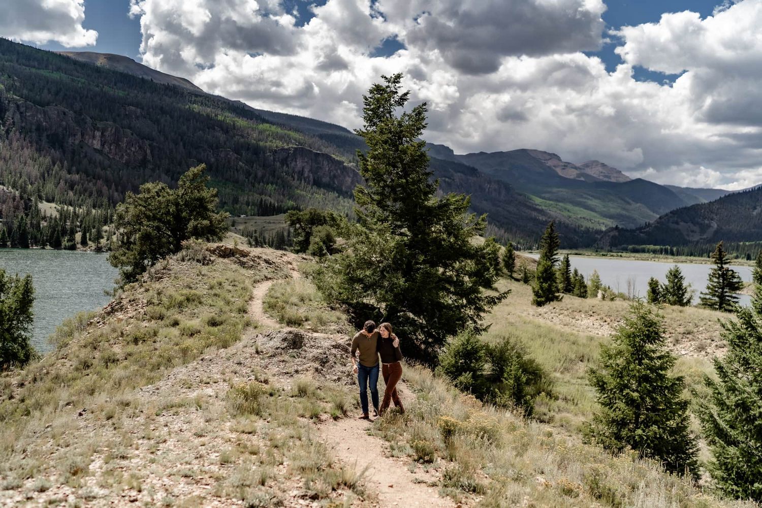Two people hiking on a mountain trail overlooking a serene lake and evergreen forest.