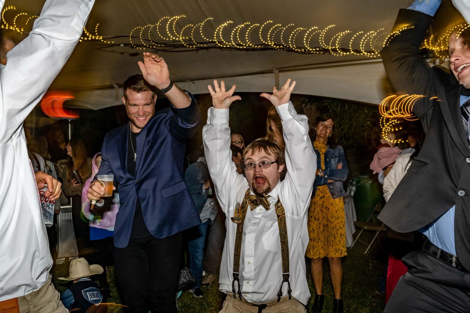 Wedding guests dancing enthusiastically with raised arms under decorative lights at reception.