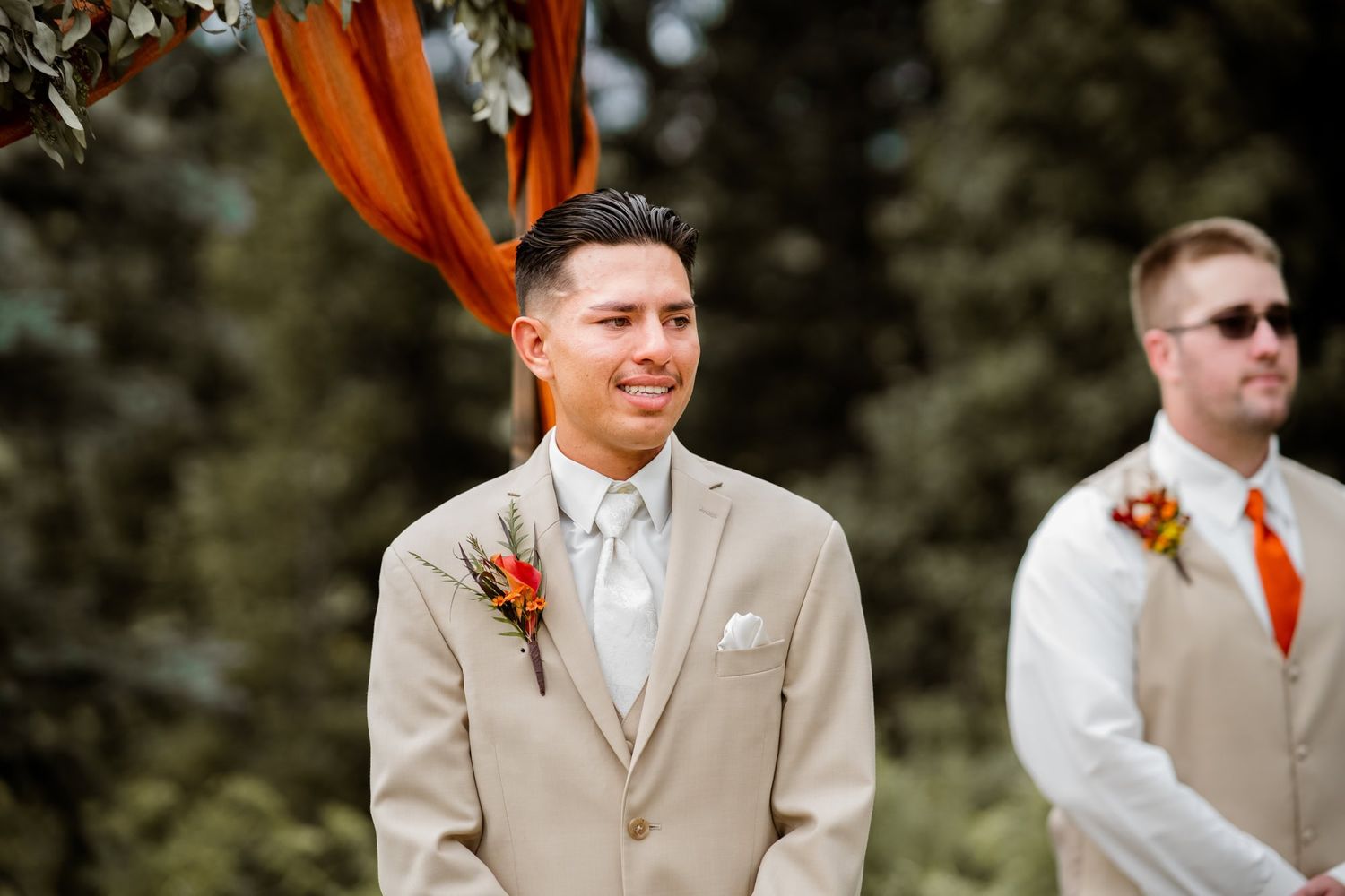 Wedding party members in beige and orange attire stand during outdoor ceremony.