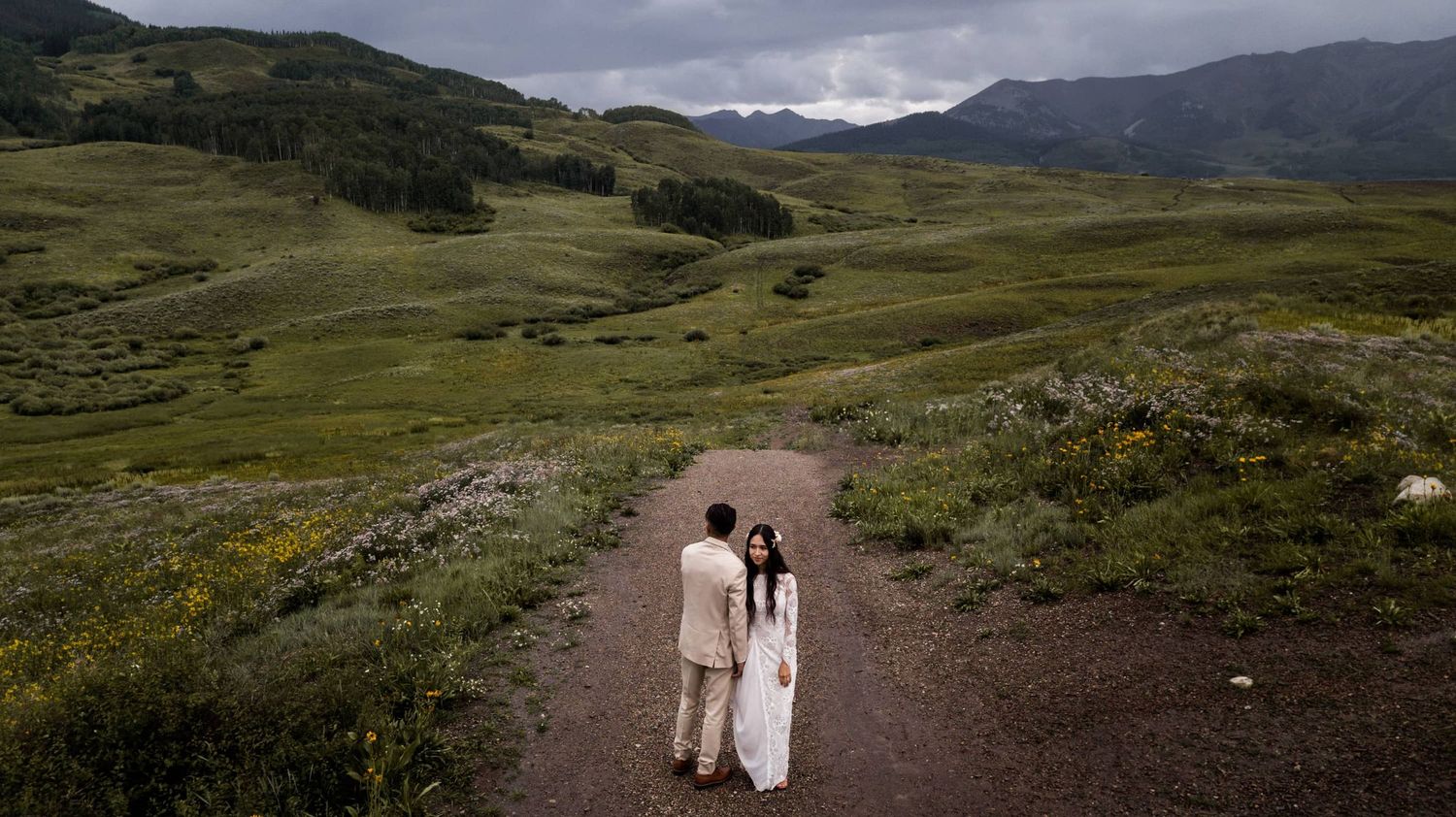 Bride and groom walk hand in hand on a dirt path through rolling green hills and mountains. Located at Mountain garden wedding venues