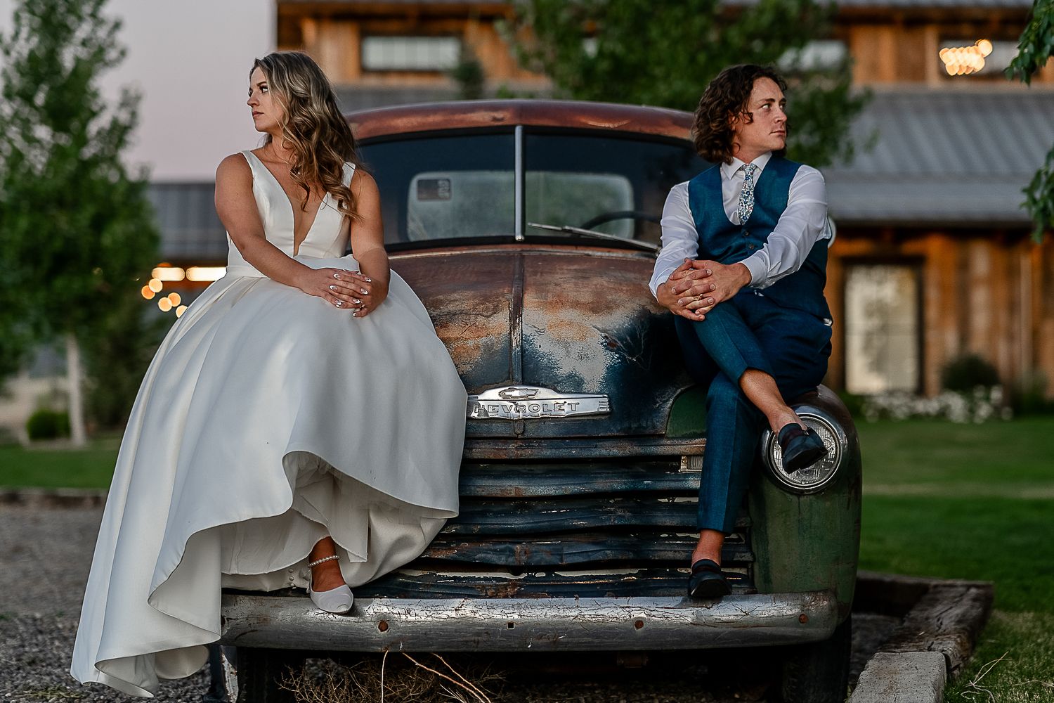 Couple poses on vintage truck at rustic wedding venue during sunset.