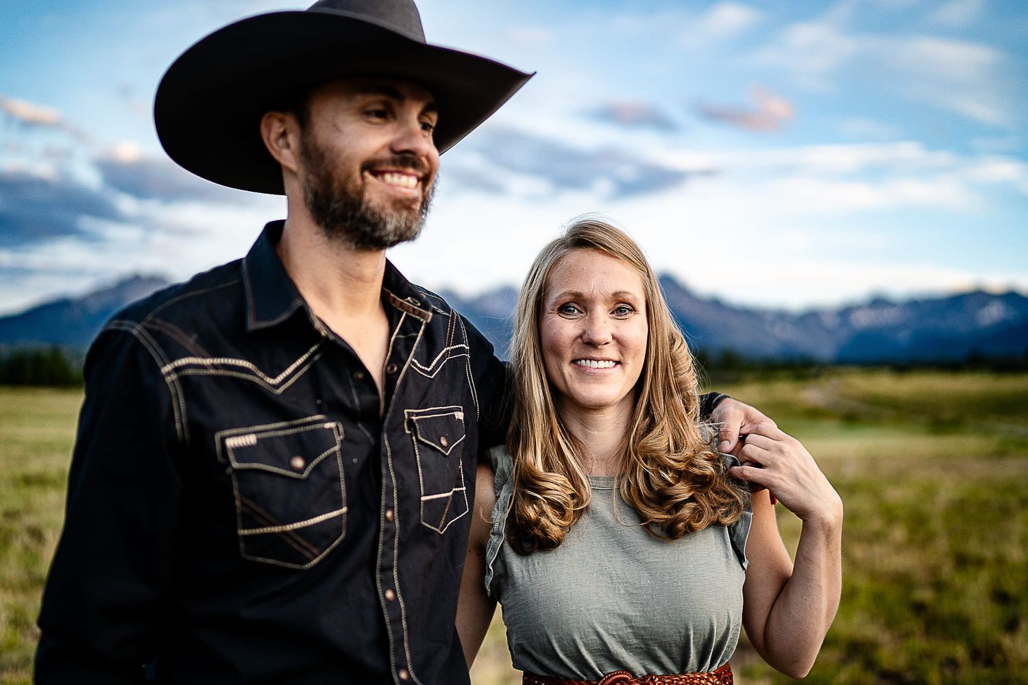 Couple in western wear smiling together in mountain meadow.