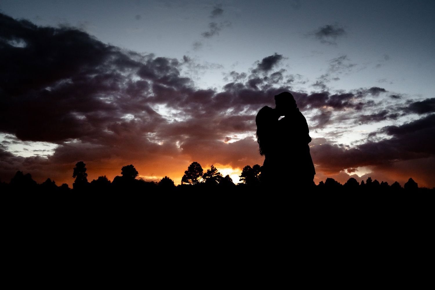 Silhouetted figures stand against dramatic orange and purple sunset sky with trees in background.