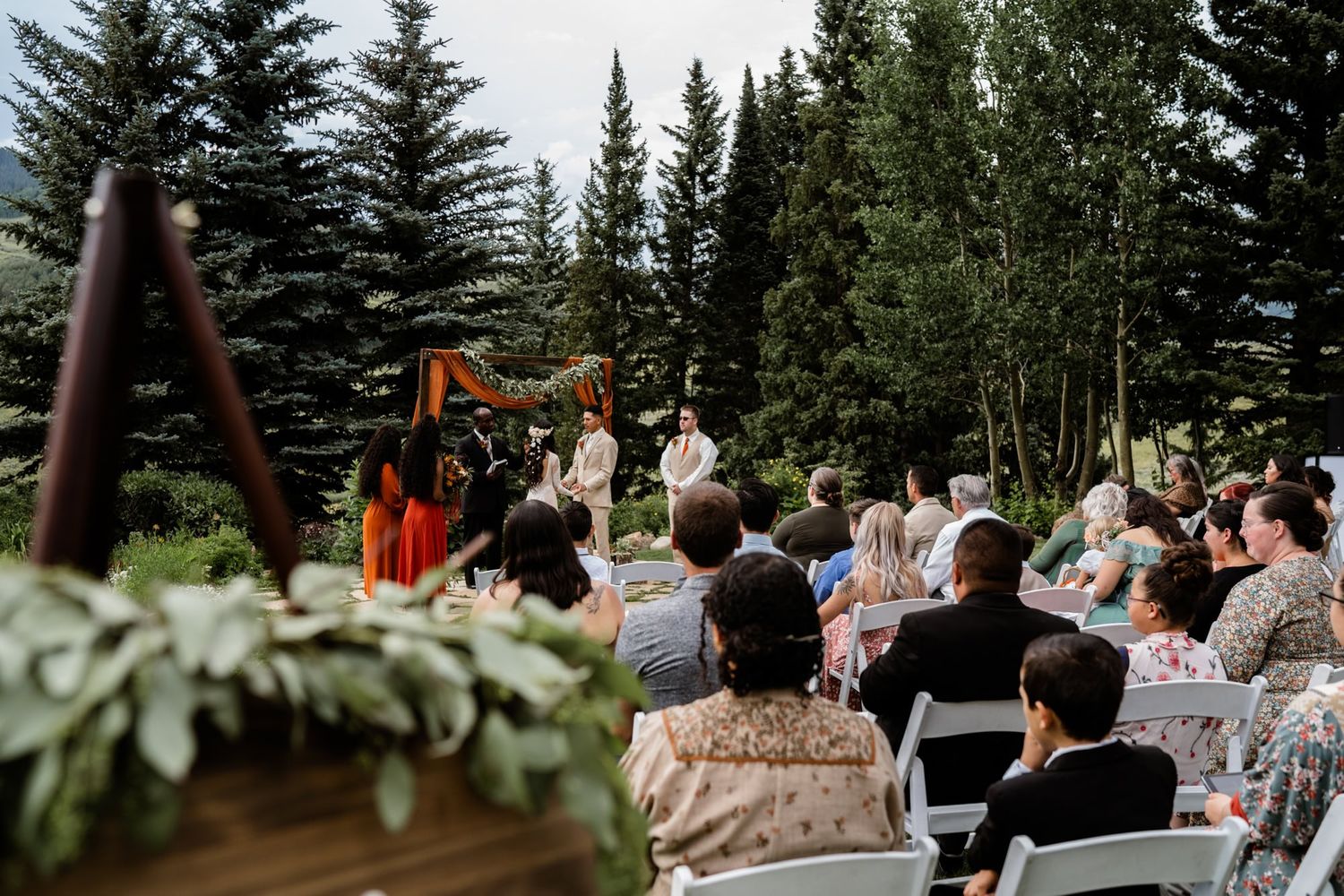 Outdoor wedding ceremony in progress with guests seated on white chairs surrounded by evergreen trees and mountain scenery.