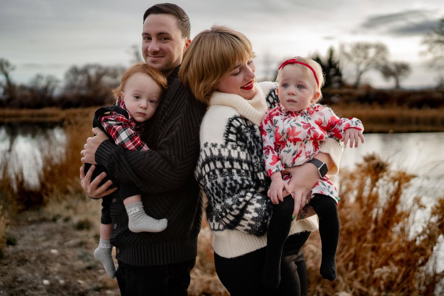 A family in winter sweaters poses together by a lake with tall grass in the background.